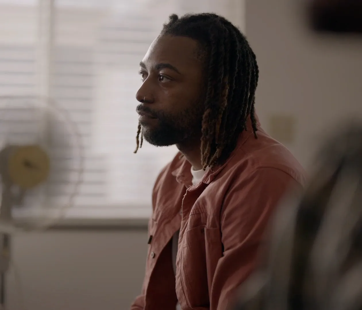 A man with locs in a pink shirt, seated indoors, looking off camera.
